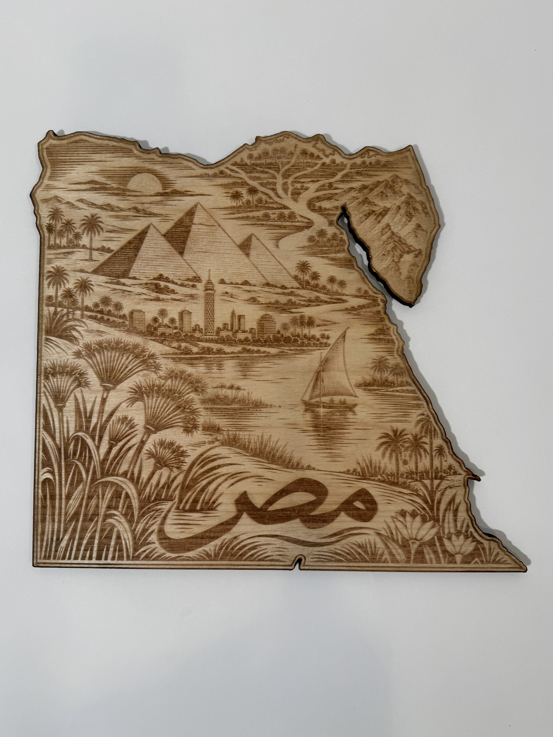 Real product photograph of Egypt wood wall art, showing pyramids, a river sailboat, skyline, palms, flowering reeds and مصر and natural wood grain.
