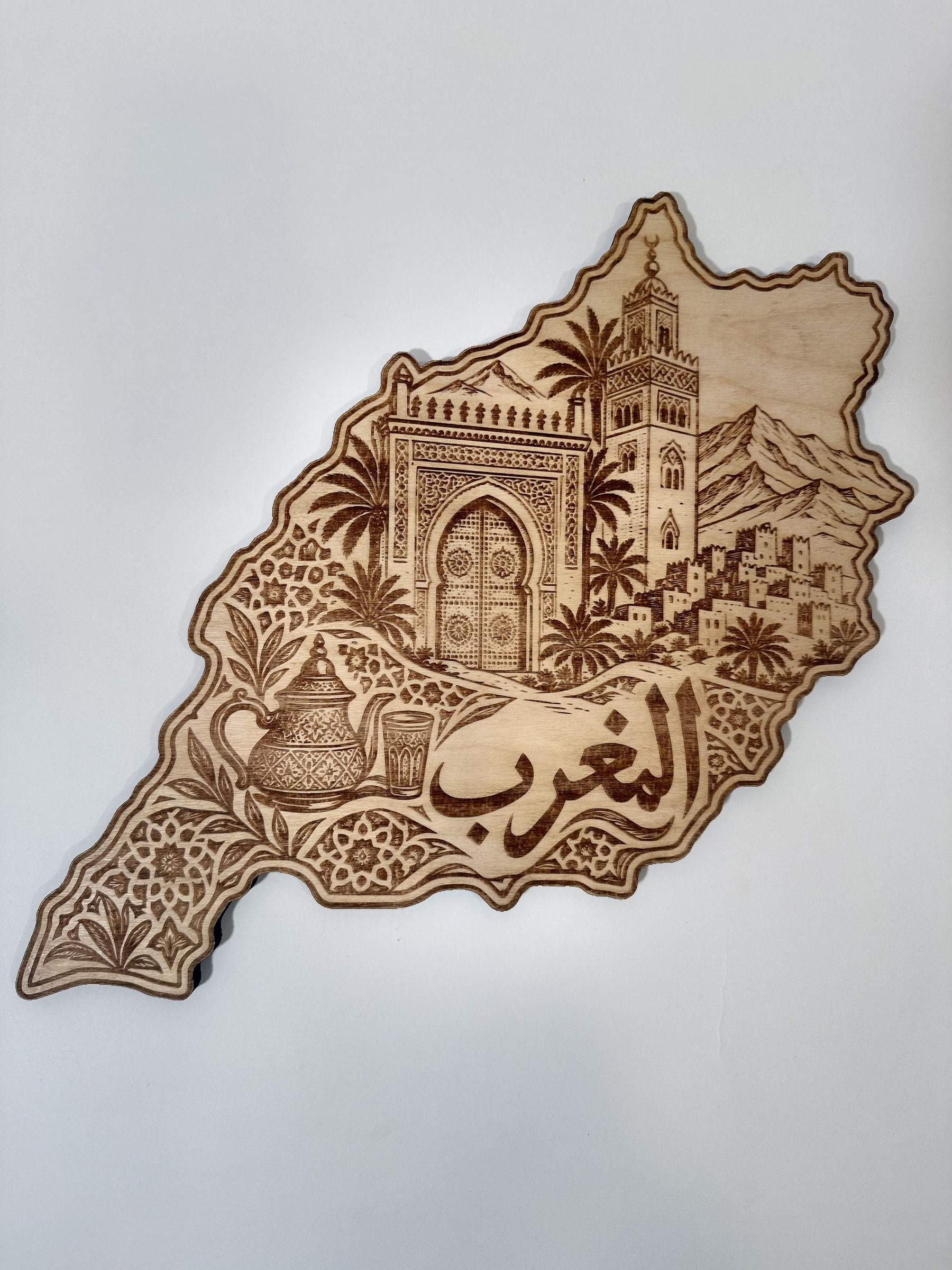 Real product photograph of Morocco wood wall art, showing an ornate gateway, minaret, mountains, palms, tea set and المغرب and natural wood grain.