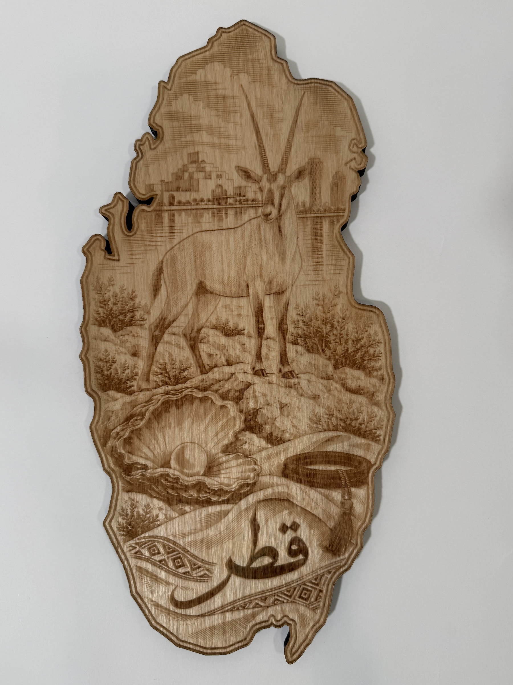 Real product photograph of Qatar wood wall art, showing an Arabian oryx, waterfront skyline, pearl shell, folded cloth and قطر and natural wood grain.