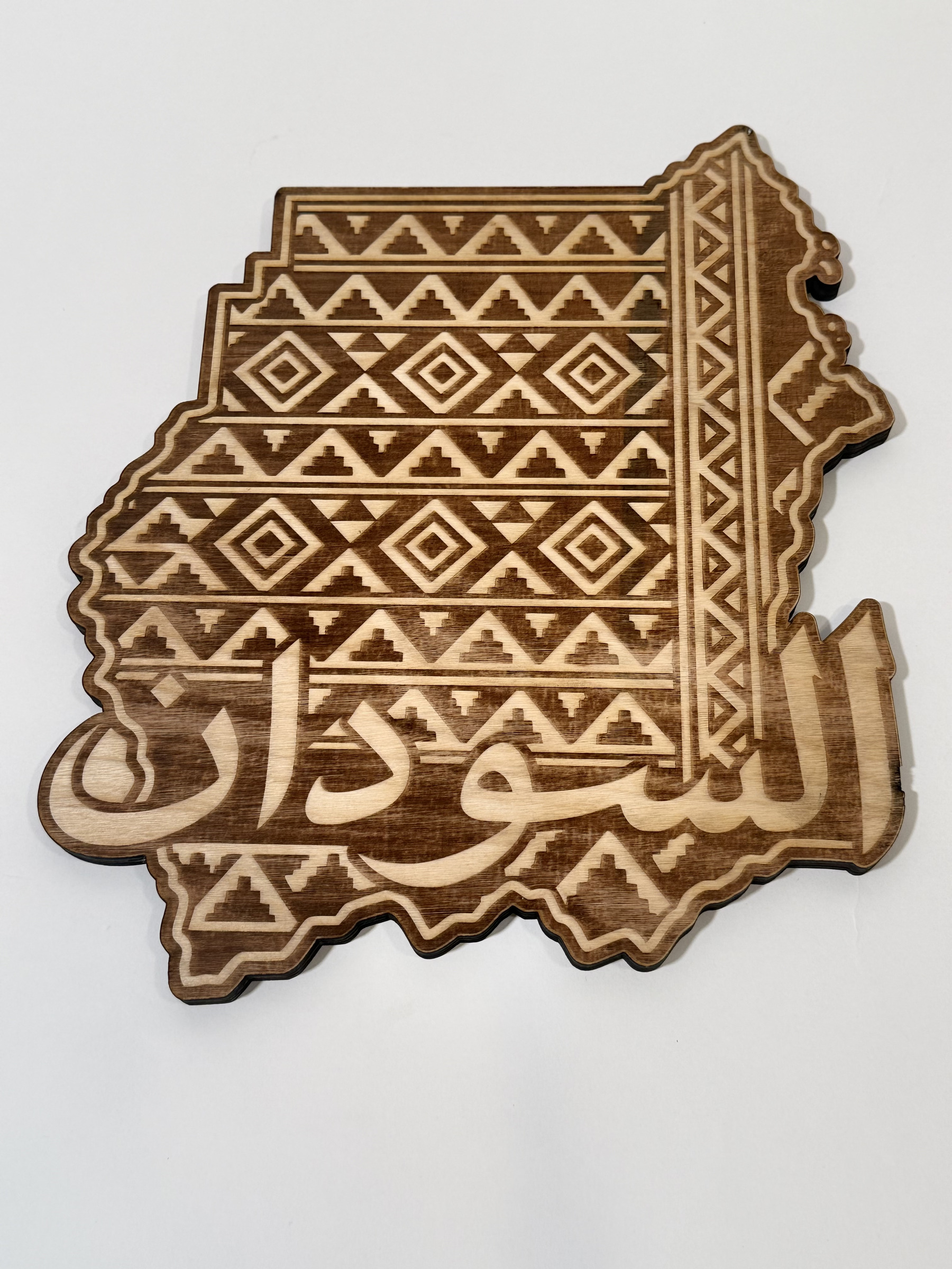 Real product photograph of Sudan wood wall art, showing stepped triangles, diamonds, geometric bands and large السودان calligraphy and natural wood grain.