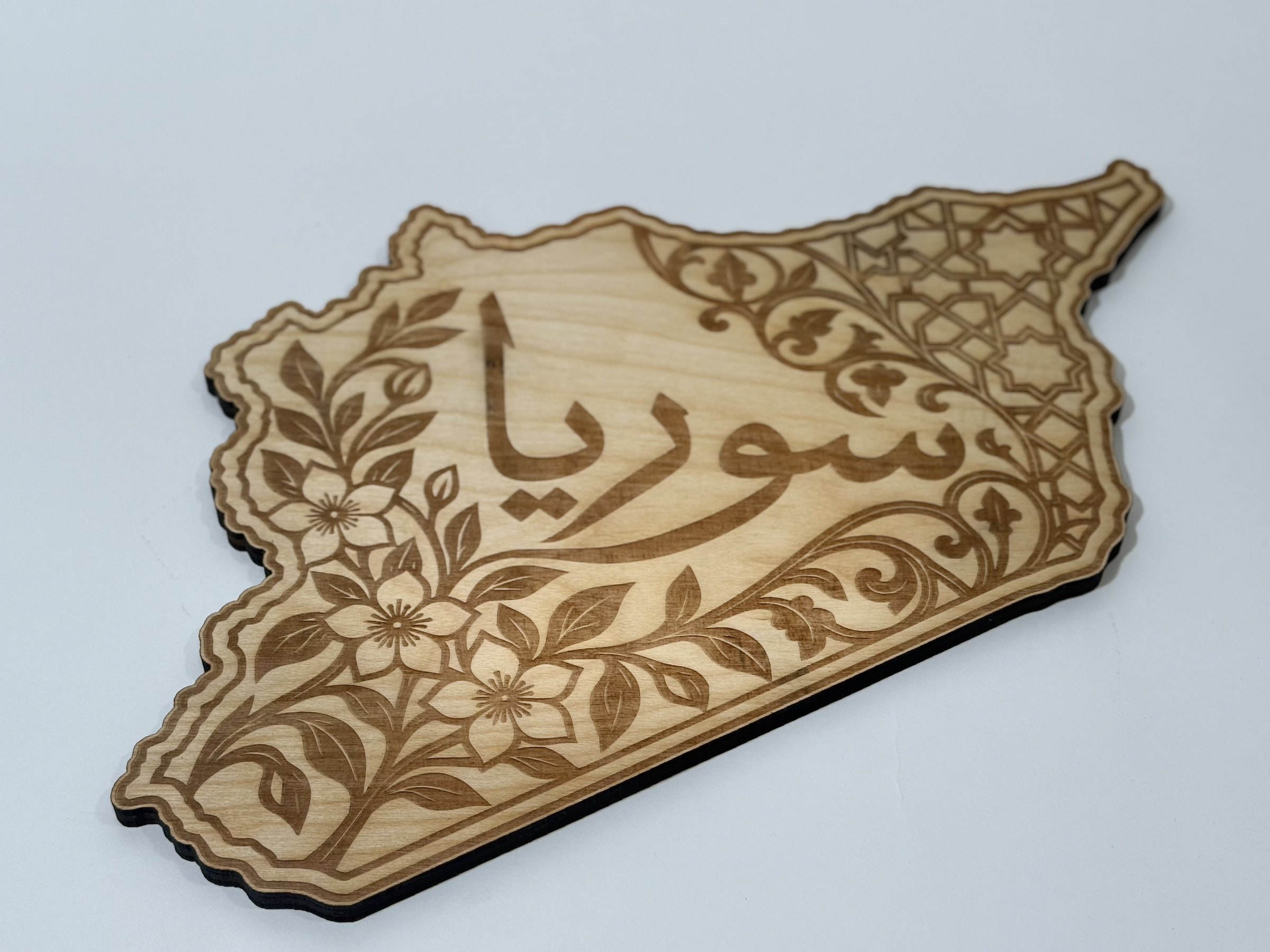 Real product photograph of Syria wood wall art, showing سوريا calligraphy framed by flowers, leaves, scrollwork and star geometry and natural wood grain.