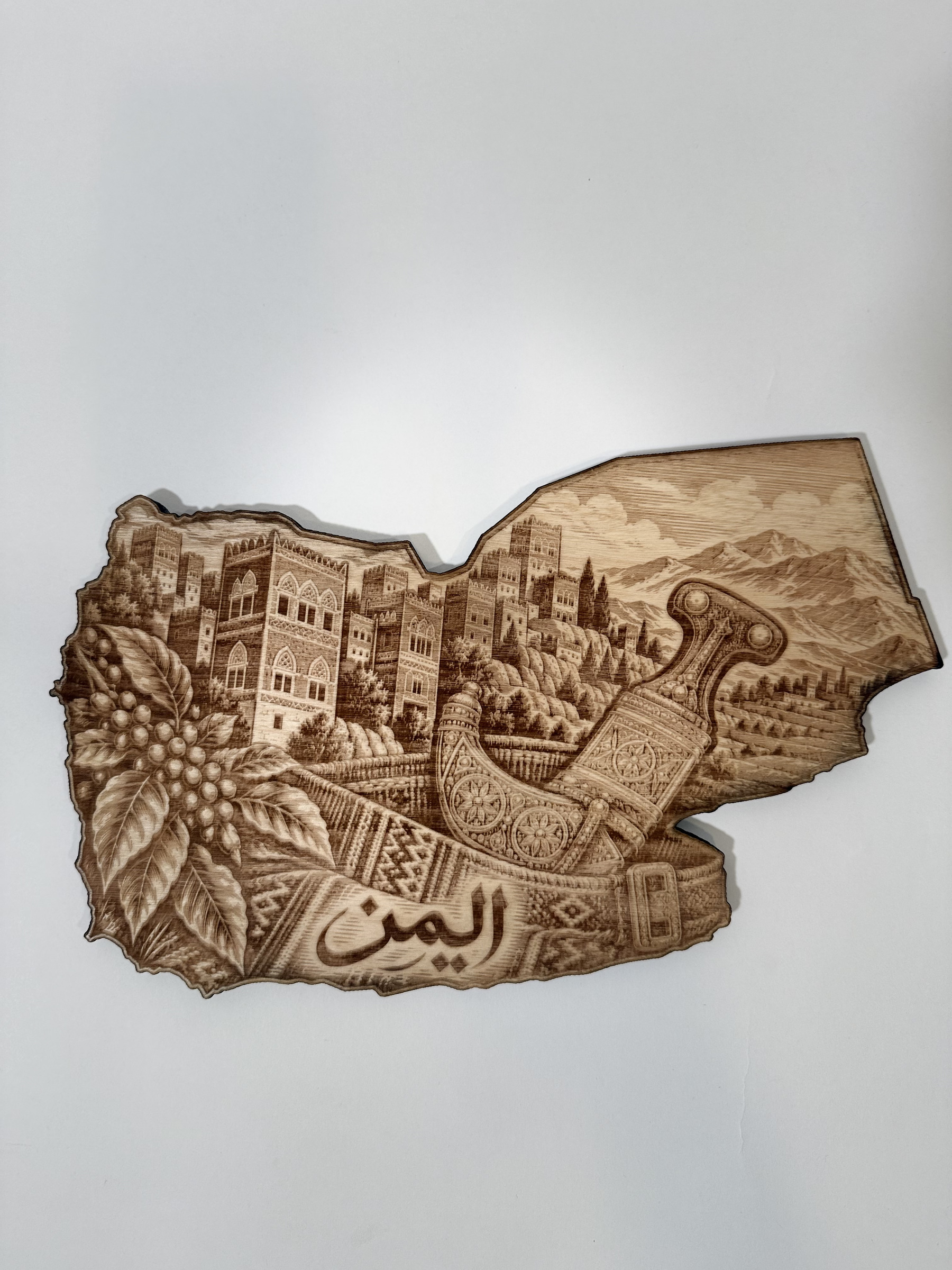 Real product photograph of Yemen wood wall art, showing ornate buildings, mountains, coffee branches, a jambiya, patterned belt and اليمن and natural wood grain.